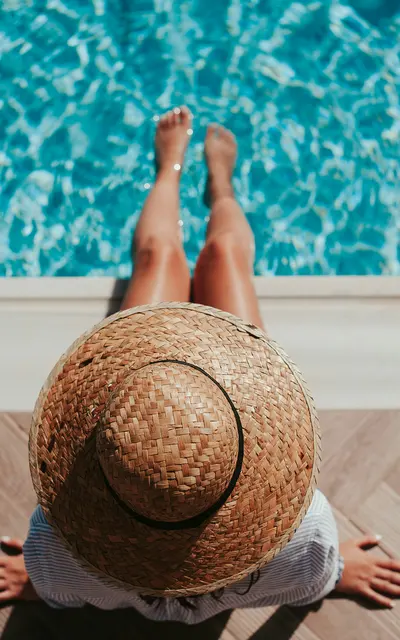 Bayshore Pines A woman in a straw hat sits by the edge of a swimming pool, with her legs dipped in the water. The pool's turquoise water reflects the sunlight, and the scene is relaxed and summery.