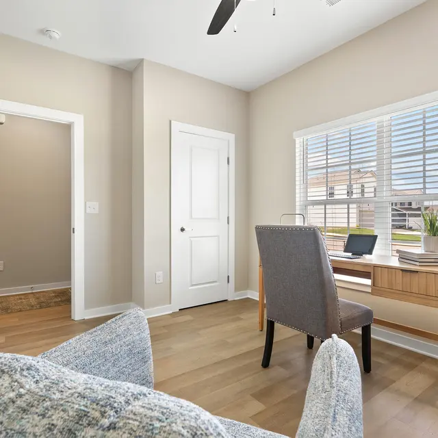 A cozy home office space featuring a wooden desk with a laptop and a small plant. There are light-colored walls, a ceiling fan, and large windows with blinds. A comfortable chair is positioned in front of the desk, and a glimpse of a couch is visible in the foreground.