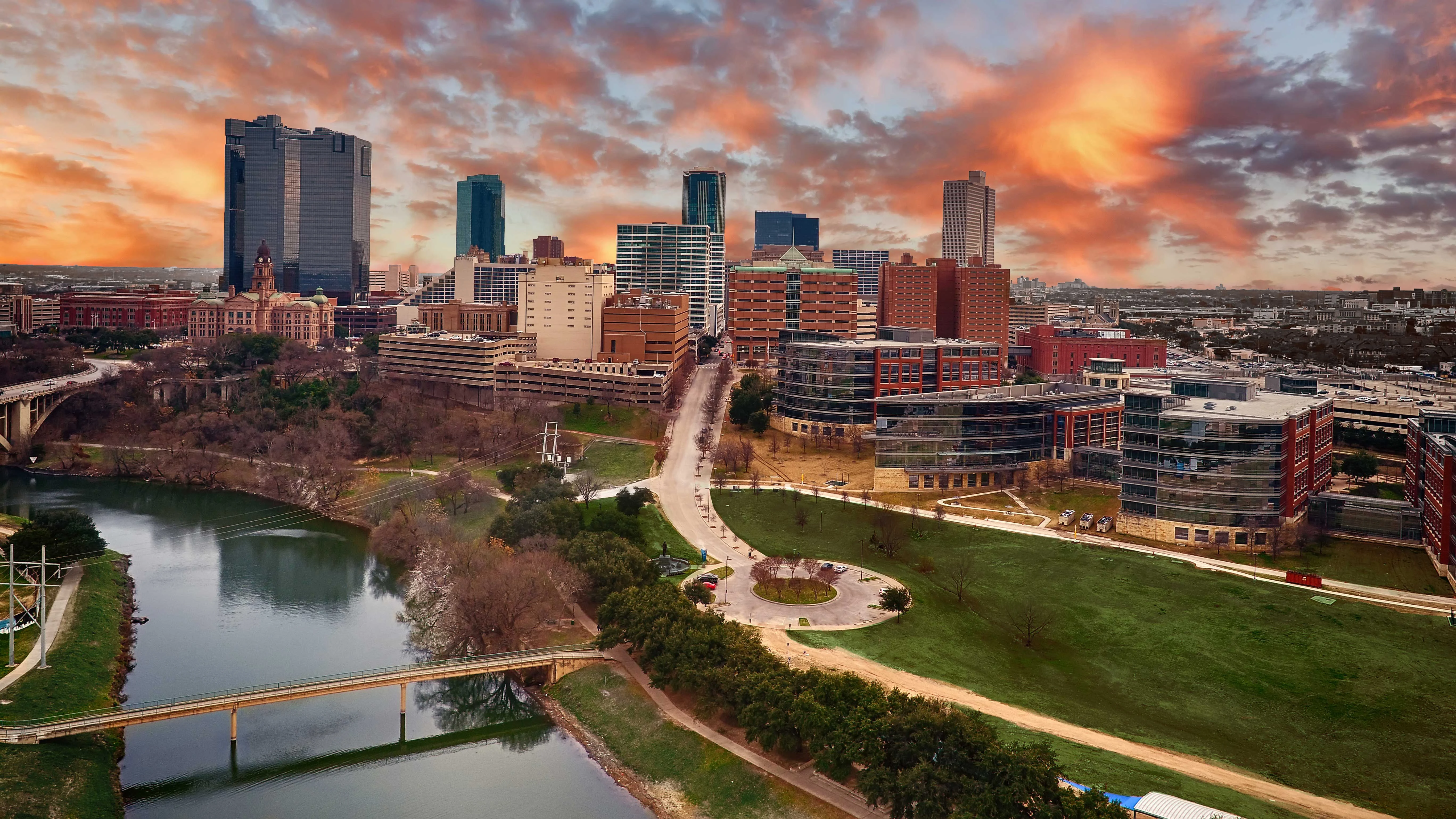 Aerial view of a modern city skyline at sunset with colorful clouds, reflecting off buildings. A river flows alongside green parks and pathways.