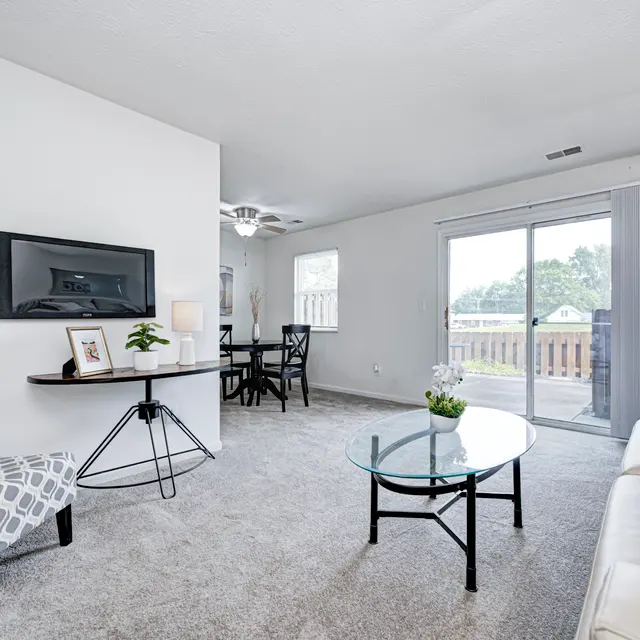 A cozy living room featuring a beige sofa, a patterned chair, a circular glass coffee table, and a television mounted on the wall. There is a sliding glass door leading out to a deck and a dining area visible in the background.