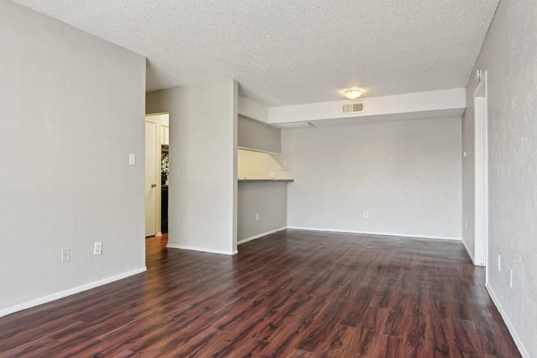 An empty living space with light gray walls and dark wood laminate flooring. The area has an open layout, leading to a kitchen space with a bar. A doorway in the background suggests access to another room.