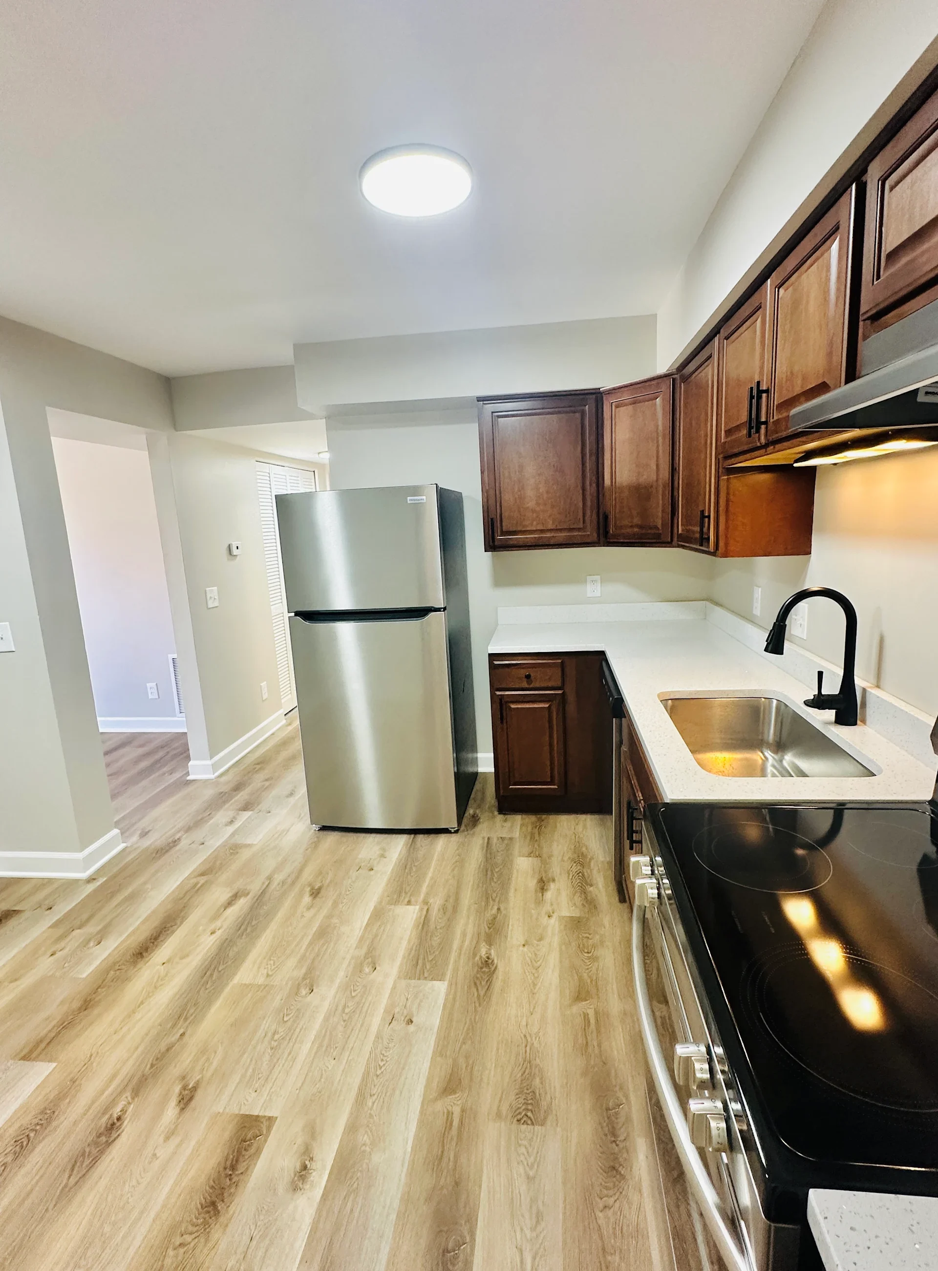 A modern kitchen featuring a stainless steel refrigerator, dark wooden cabinets, and a black countertop. The kitchen has a spacious layout with laminate flooring and a ceiling light.