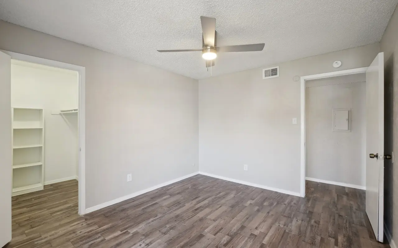 A spacious empty room with a ceiling fan, light-colored walls, and wood-style flooring. A doorway leads to a closet on one side, while another door leads outside the room.