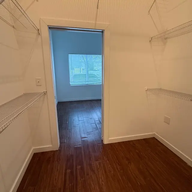 An empty closet featuring wire shelves on the walls, with a doorway leading to a room visible in the background. The floor is wooden and there is a window that shows trees outside.