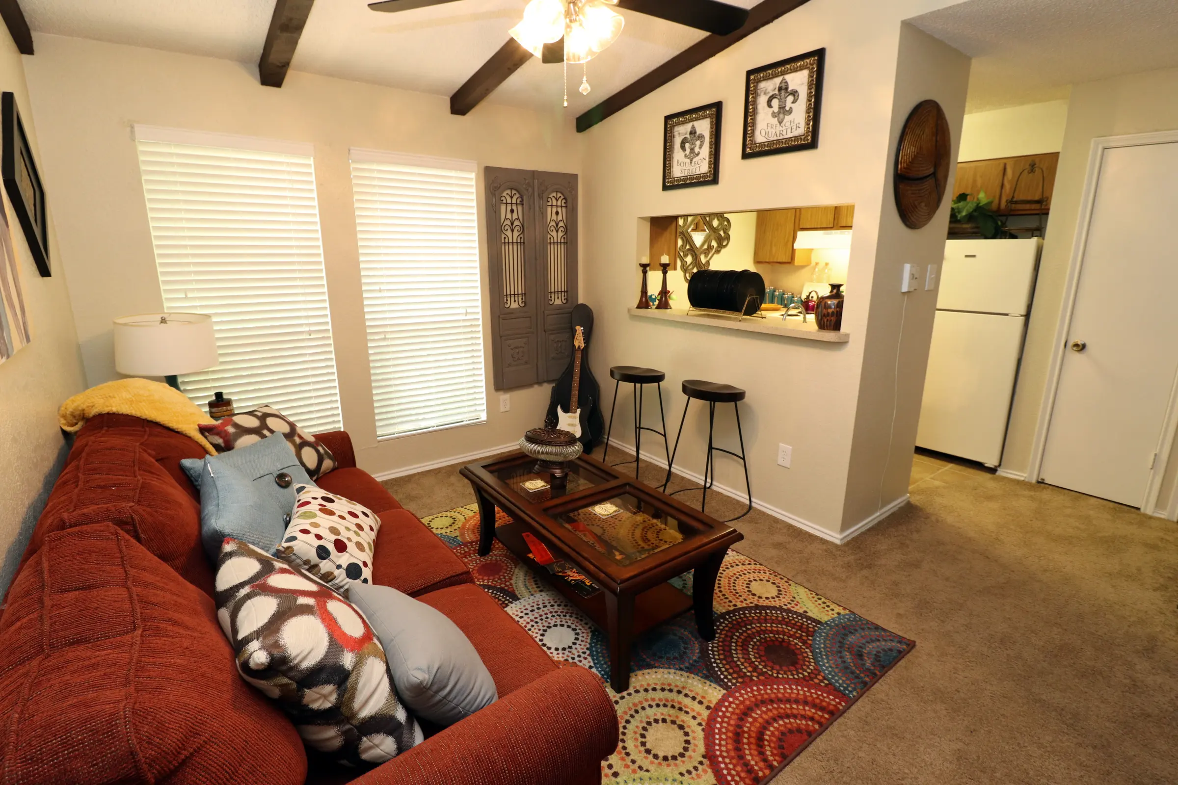 Alexis Park A cozy living room featuring a red sofa adorned with colorful pillows, a coffee table, and a patterned area rug. There are blinds on the windows, a ceiling fan, and a small bar with stools connecting to a kitchen area.