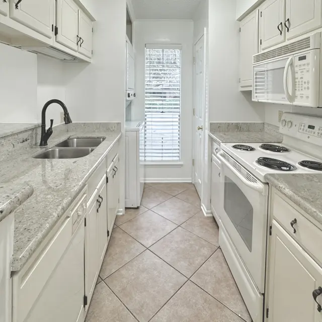 A narrow kitchen view featuring light-colored cabinets, granite countertops, and modern appliances including a stove, microwave, and sink.
