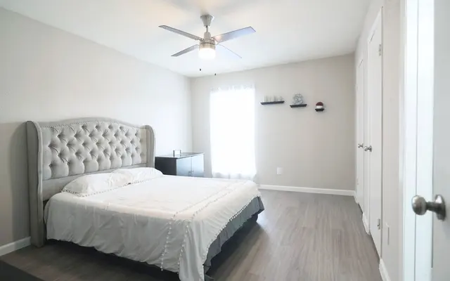 A modern bedroom featuring a light gray tufted bed with a white bedspread, a nightstand, and a ceiling fan. Natural light enters through a window with sheer curtains.
