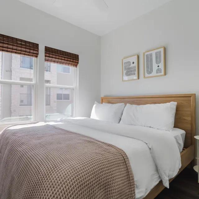 A cozy modern bedroom featuring a wooden bed with white and beige bedding, small bedside table, and two windows with bamboo blinds.