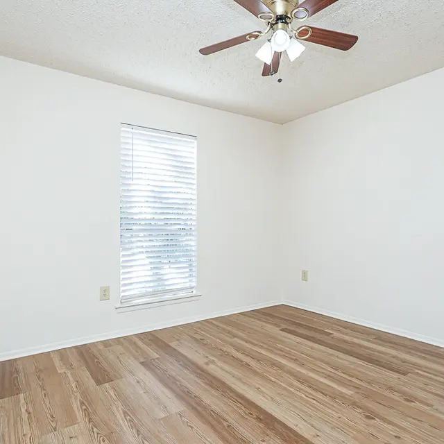 A vacant room with light wooden flooring, white walls, and a ceiling fan with lights. A window with blinds allows natural light to enter the space.