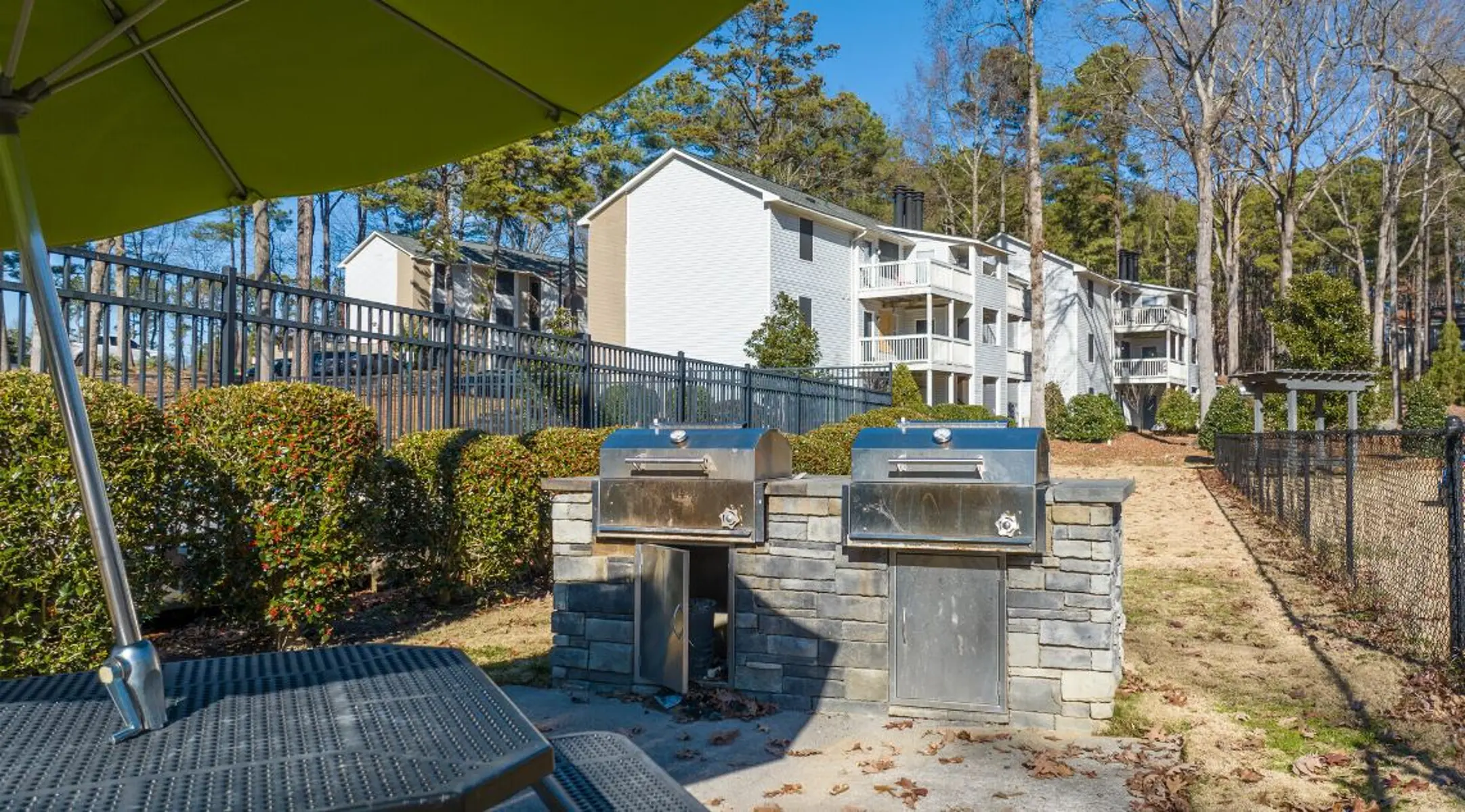 Grilling area with two metal grills, surrounded by bushes and a few trees, with a view of an apartment building in the background.
