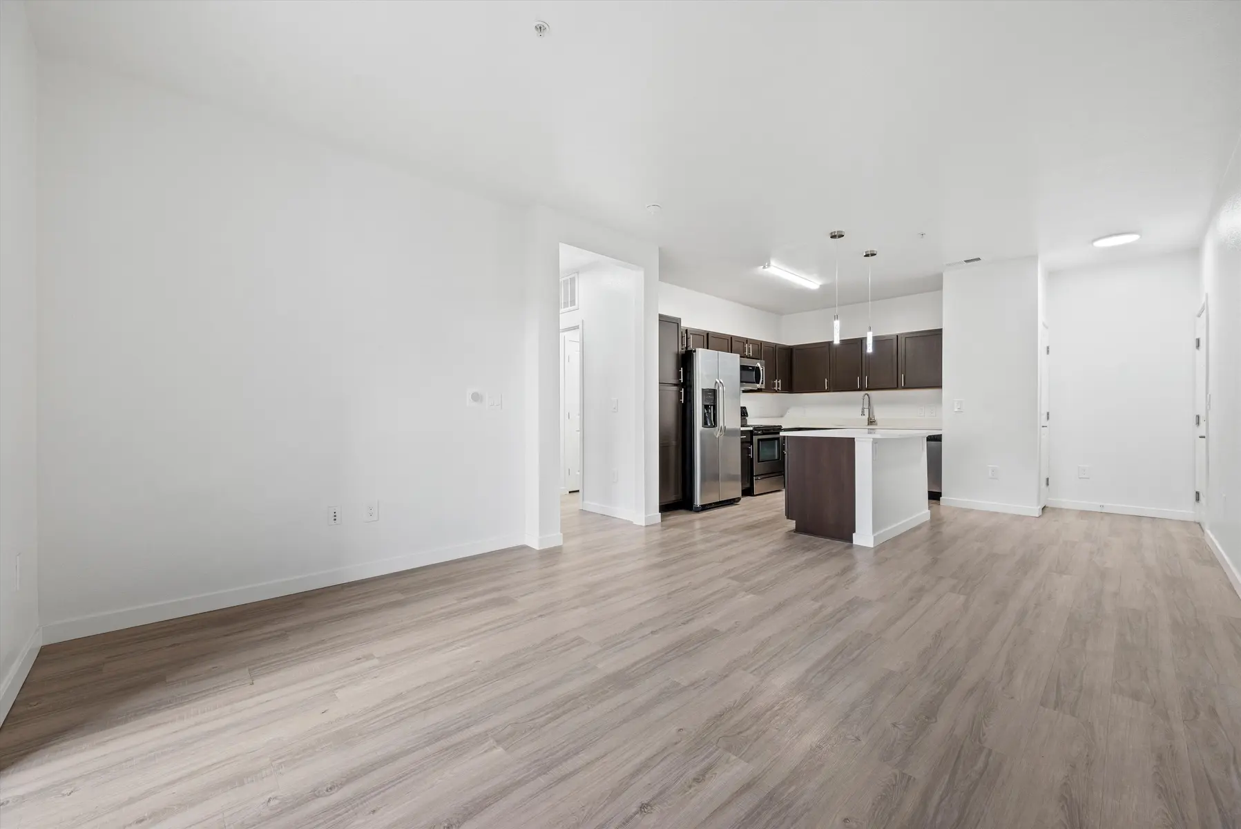 A modern apartment interior featuring an open layout with a kitchen island and stainless steel appliances. The space has light-colored walls and wooden flooring.