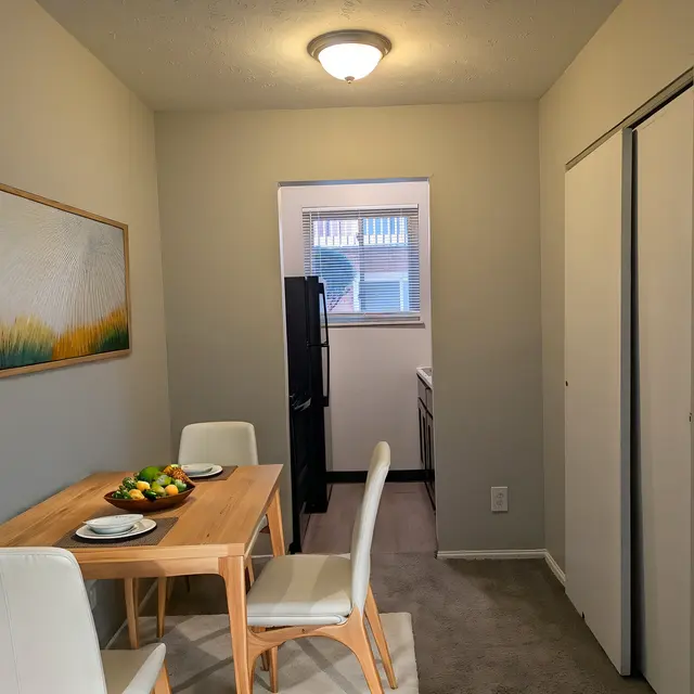 A cozy dining area featuring a wooden table with white chairs, adorned with a bowl of fruit and two place settings. A doorway leads to a kitchen area in the background, and a painting of a landscape is hung on the wall.