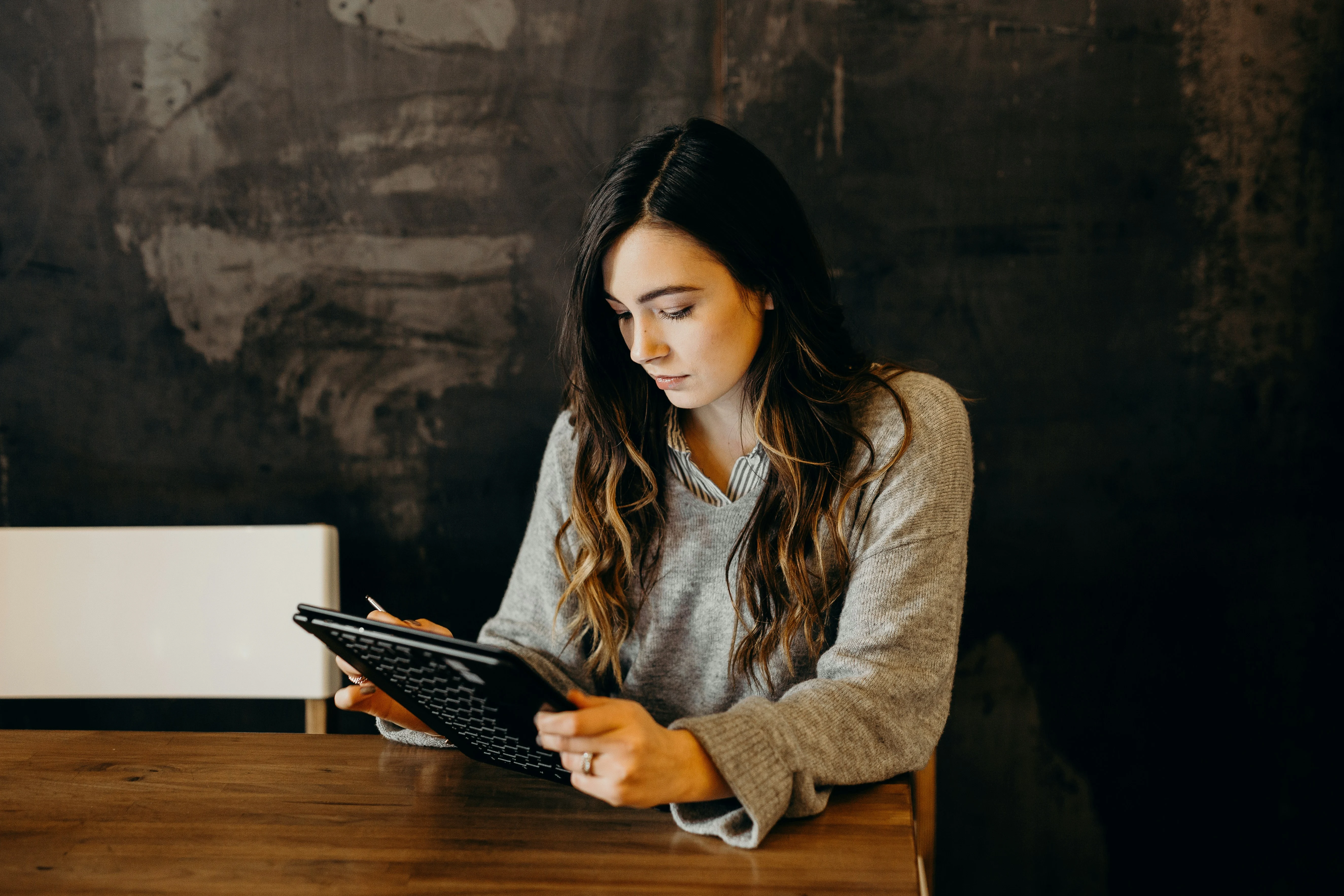 A young woman sitting at a wooden table, looking at a tablet device. She has long, wavy hair and is wearing a light gray oversized sweater. The background features a dark wall with a textured surface.
