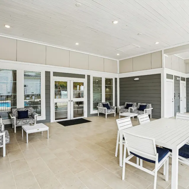 A modern outdoor lounge area with white furniture, blue cushions, and large windows overlooking a pool. Bright and spacious setting.