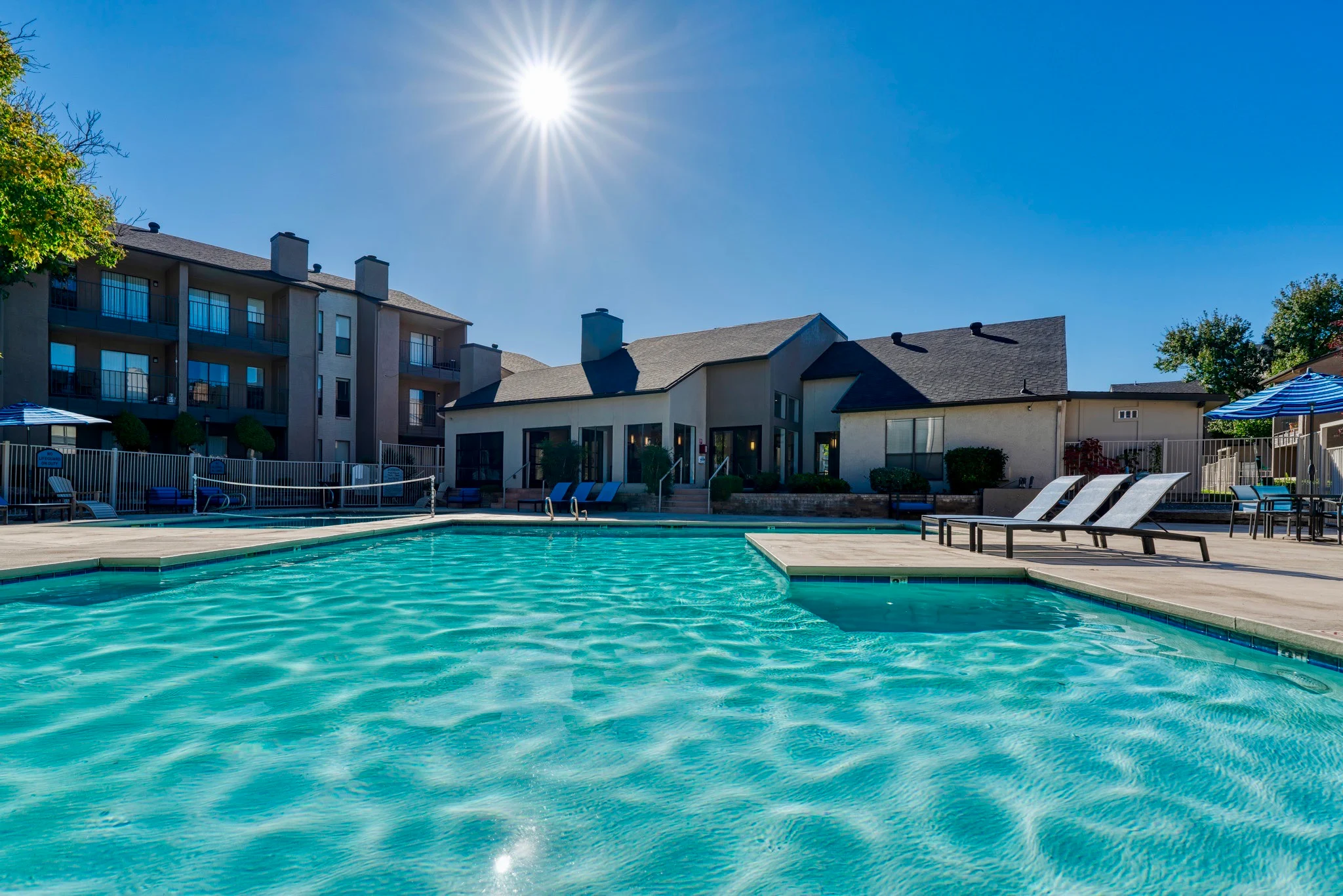 Sunlit Pool Area at Apartment Complex An outdoor swimming pool at an apartment complex on a sunny day, with lounge chairs by the pool and a building in the background.