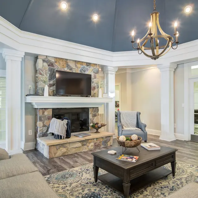 A cozy living room featuring a stone fireplace with a TV mounted above it, elegant chandelier lighting, and comfortable seating arrangements including a sofa and armchair. The room is painted in light tones with a dark blue ceiling, complemented by large windows for natural light.