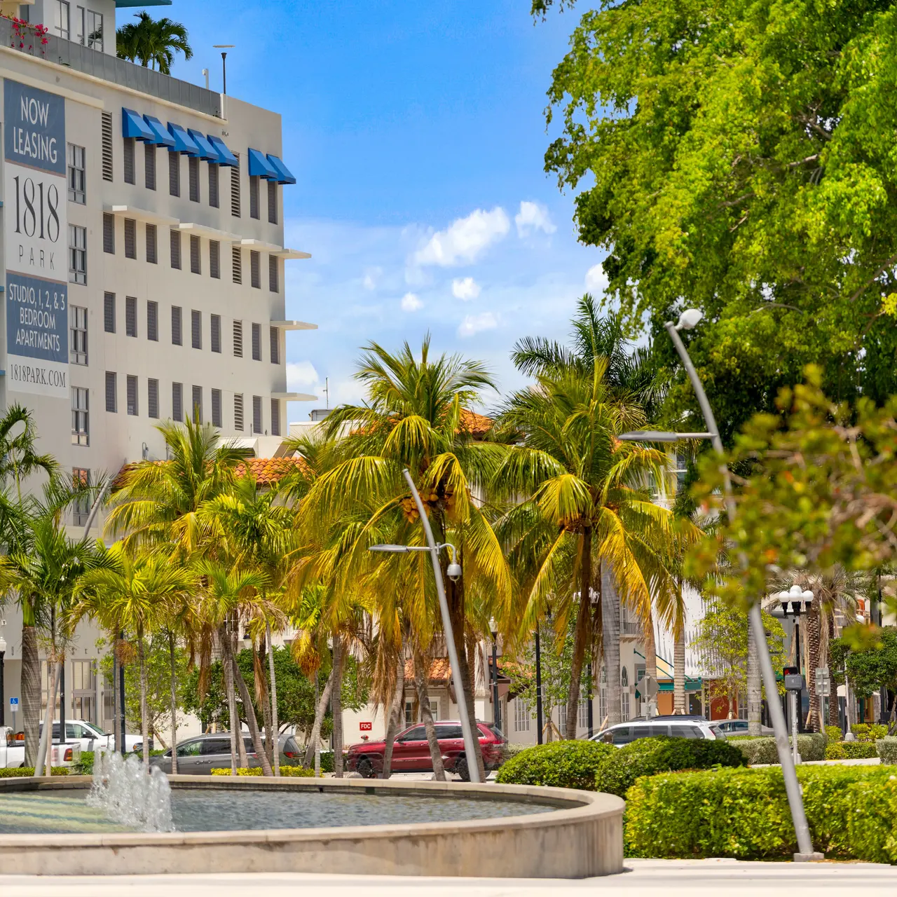 A park scene featuring a fountain, surrounded by palm trees and modern buildings in the background. Bright blue sky with scattered clouds adds to the vibrant atmosphere.
