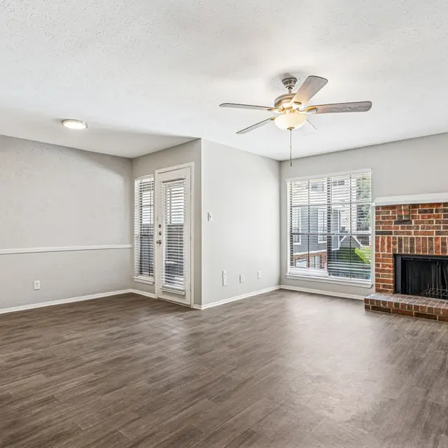 Spacious living room with wooden flooring, a ceiling fan, a brick fireplace, and large windows.