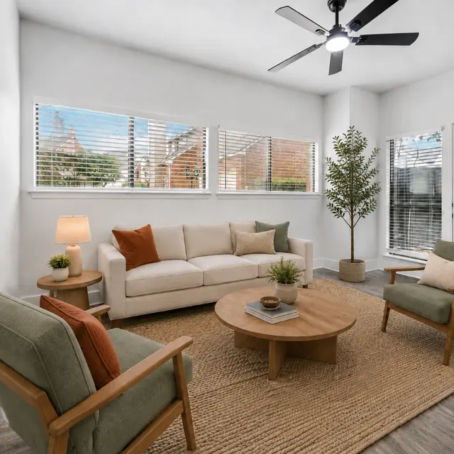 A modern living room featuring a light-colored sofa, two green armchairs, a round wooden coffee table, and a textured area rug. There are large windows with blinds, a potted plant, and a floor lamp present.
