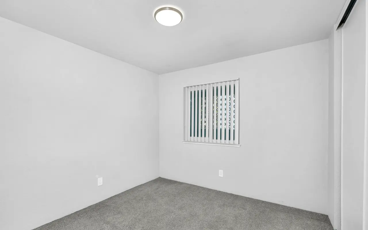An empty room with light gray carpet and walls. There is a window with vertical blinds on one wall and a ceiling light fixture.