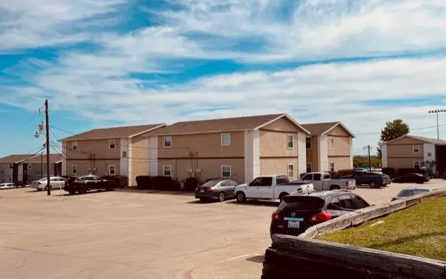 View of an apartment complex with several vehicles parked in the lot, under a blue sky with clouds.