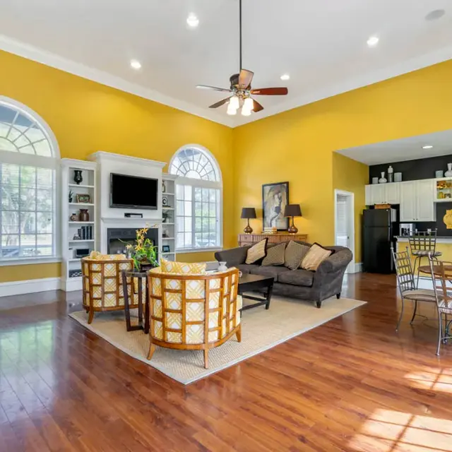 Bright and spacious living room featuring high ceilings, large windows, and wooden flooring. The space includes a seating area with couches and chairs, a coffee table, and a dining area with a metal table and chairs, all set against a vibrant yellow wall.