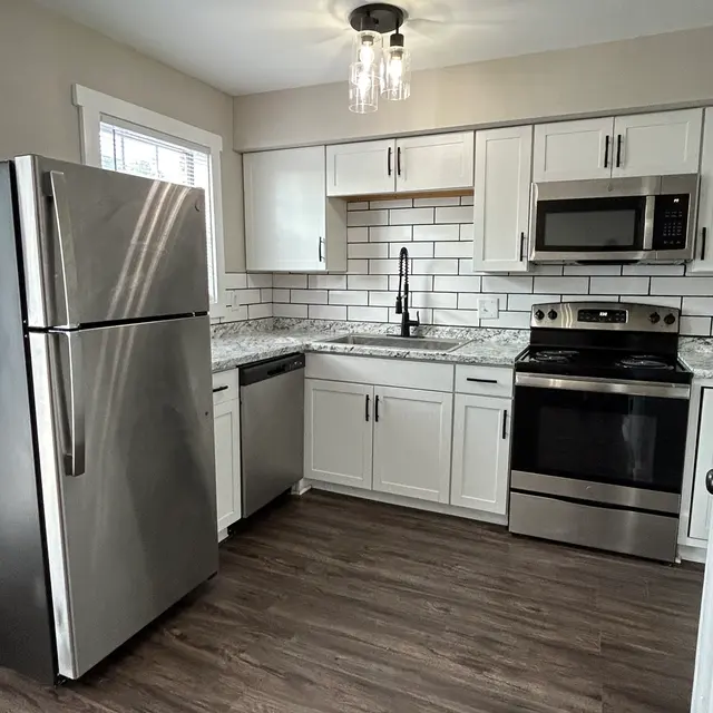 A modern kitchen featuring stainless steel appliances, including a refrigerator, stove, and microwave, with granite countertops and white cabinetry. The backsplash is made of white tiles. There is a natural wood floor and a stylish overhead light fixture.