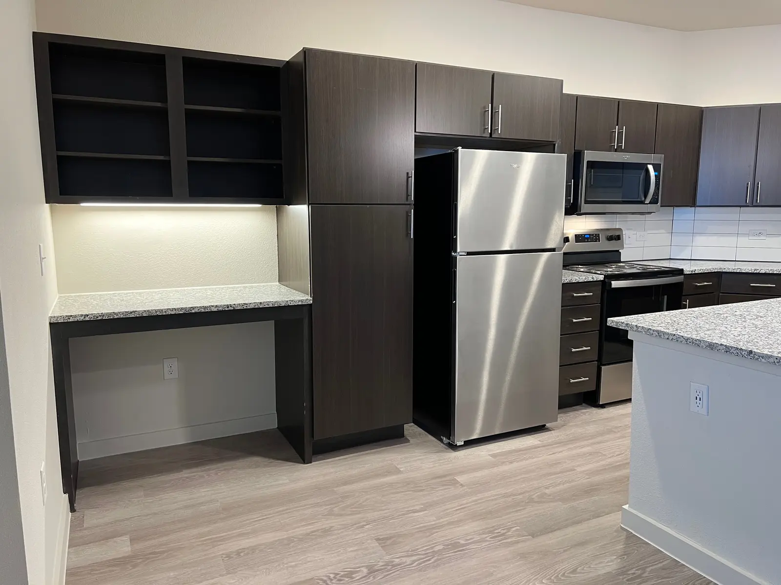 Modern Kitchen Design A modern kitchen featuring dark cabinetry and light countertops. There is a stainless steel refrigerator and a built-in microwave. The space includes a small desk area and hardwood flooring.
