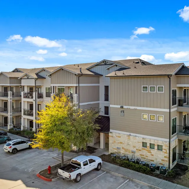 An exterior view of a modern three-story apartment complex with balconies, surrounded by trees and parked cars.