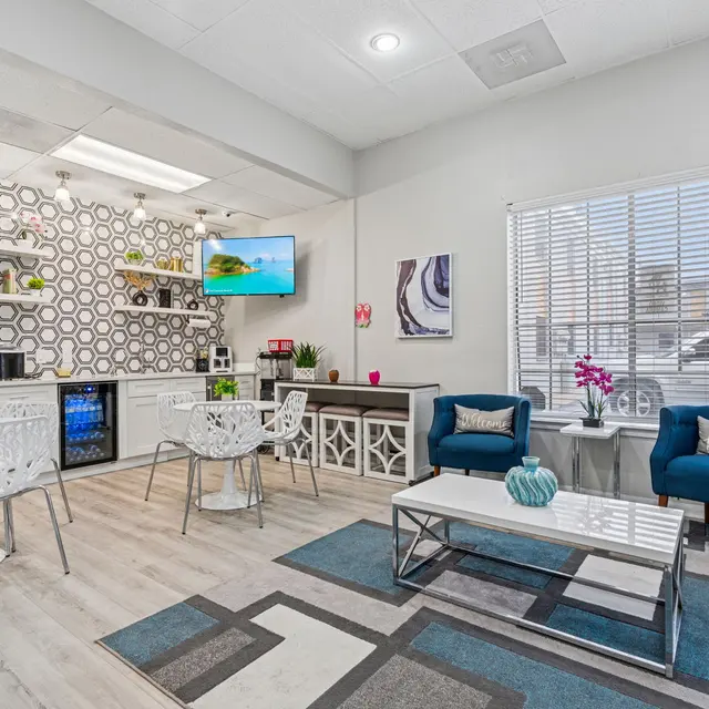 A modern kitchen interior featuring a geometric patterned wall, white furniture, blue accent chairs, and an open layout with a window allowing natural light.
