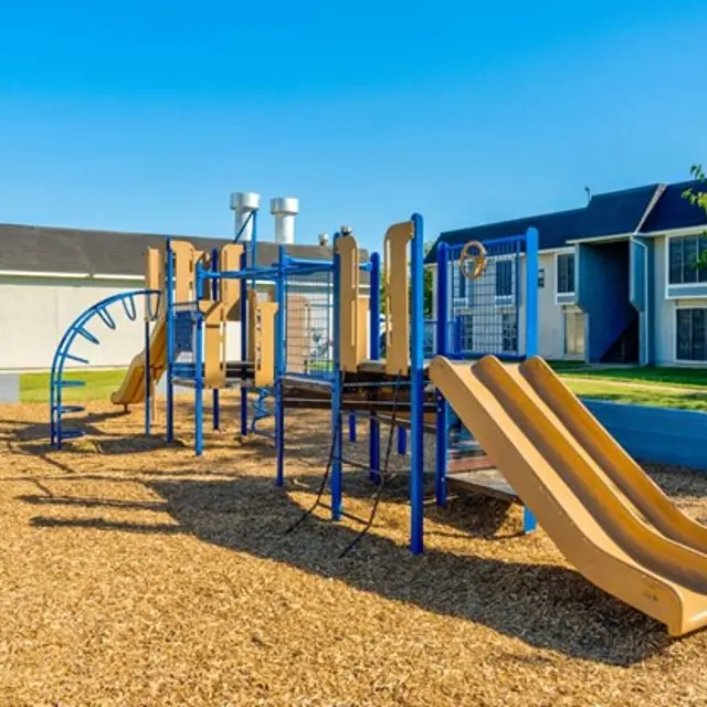 A playground featuring slides and play structures set in a sandy area with surrounding grass and buildings in the background.