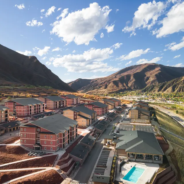 Aerial view of a housing development nestled in a mountainous area, featuring several multi-story buildings, some with pools, surrounded by colorful fall foliage and distant mountains.