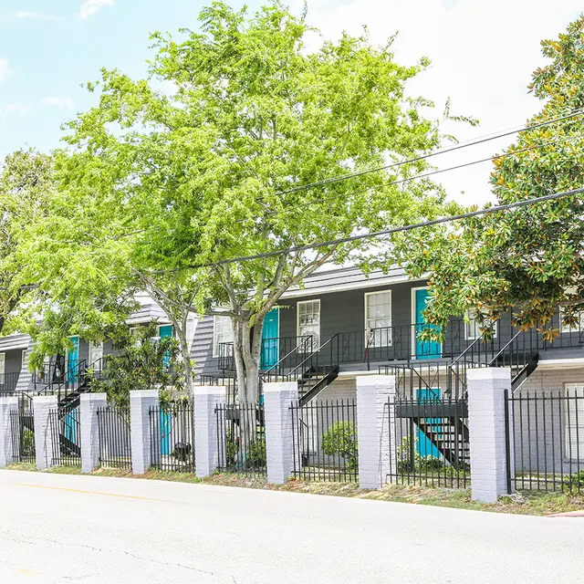 Modern Apartment Complex A view of a modern apartment complex with a row of units, surrounded by green trees and a black fence. The units feature bright blue doors and balconies.