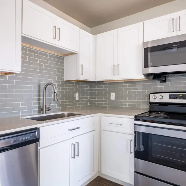 A modern kitchen with white cabinets, stainless steel appliances, and gray tile backsplash. The countertop is light-colored and features a sink. The kitchen includes a dishwasher and an oven.