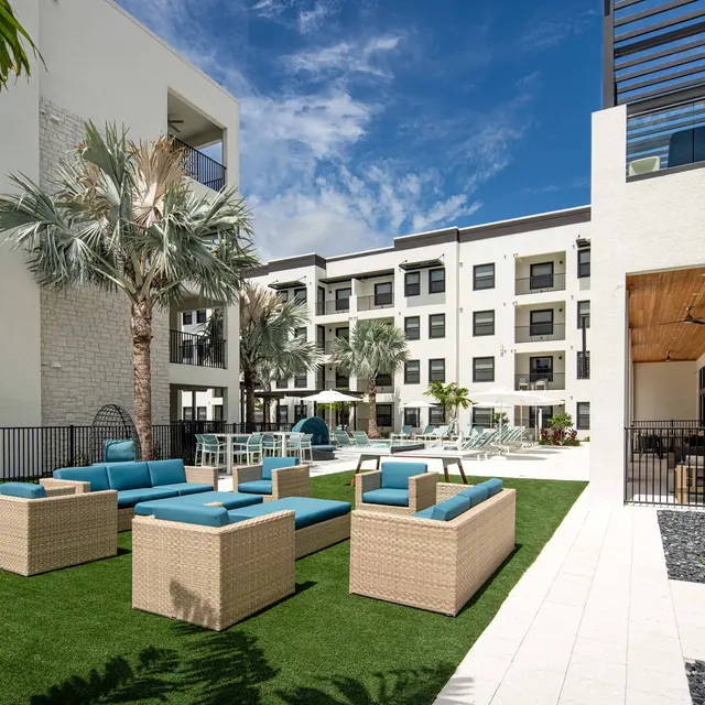 A modern courtyard area with comfortable seating, landscaped greenery, and palm trees surrounding a multi-story building.