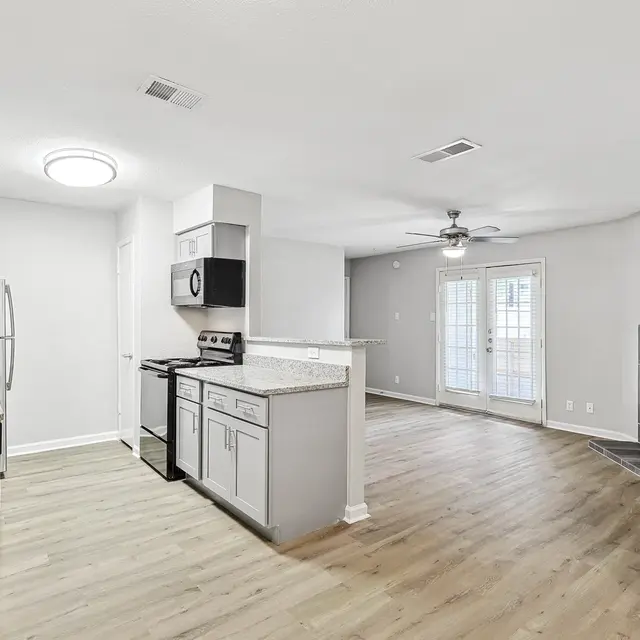 A modern apartment interior showcasing an open kitchen with granite countertops and stainless-steel appliances, leading into a living area with a ceiling fan and a fireplace.