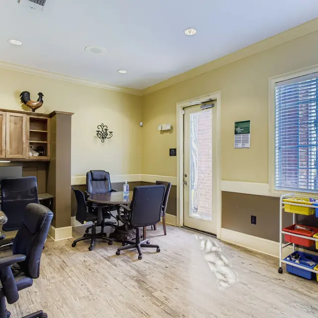 A modern office interior featuring a wooden desk with black chairs, a large window with blinds, and colorful storage bins against the wall.