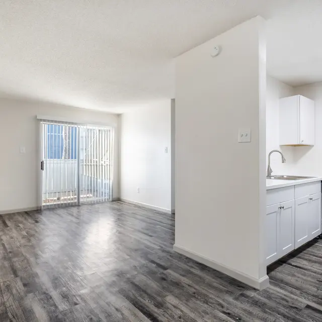 A spacious, empty apartment with wooden floors, a kitchen area on the right, and large sliding glass doors leading to a balcony on the left.