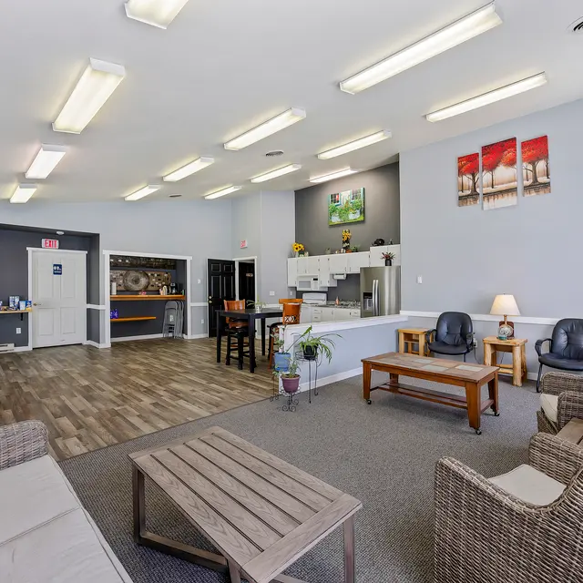 Community Area Overview A spacious community area featuring comfortable seating, a wooden table, and a kitchenette. Bright fluorescent lights illuminate the room, which has neutral-colored walls and a cozy ambiance.