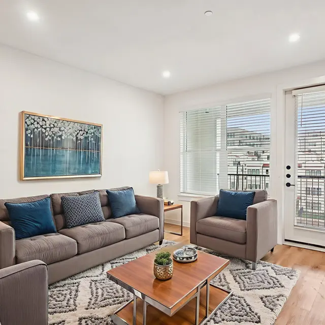 A stylish living room featuring a gray sofa and armchairs, decorated with blue cushions, a coffee table, and a large piece of artwork on the wall. There are large windows with blinds allowing natural light to fill the space and a sliding door leading to a balcony.