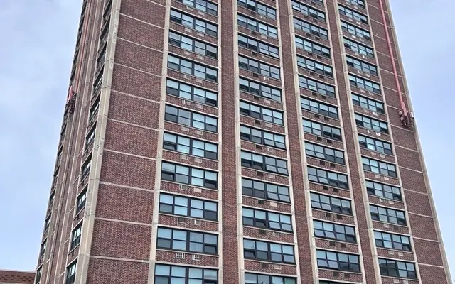 Urban High-Rise Apartment Building A tall brick apartment building with multiple windows and a small entrance area, surrounded by greenery and a stop sign in the foreground.
