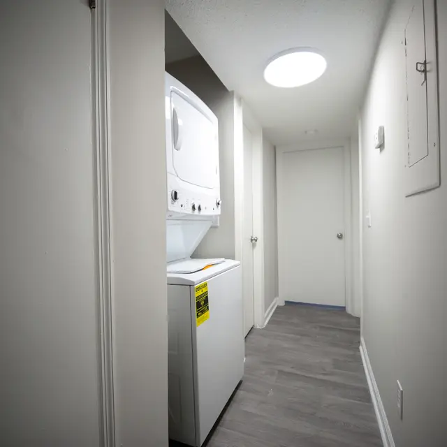 View of a hallway featuring a stacked washer and dryer, with an illuminated ceiling light, beige walls, and a door at the end of the hallway.
