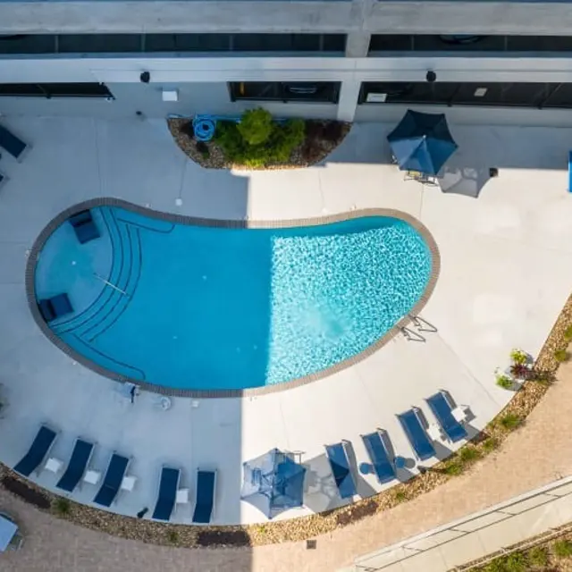 Aerial view of a modern pool area featuring a curved swimming pool surrounded by lounge chairs and umbrellas, set in a manicured courtyard.