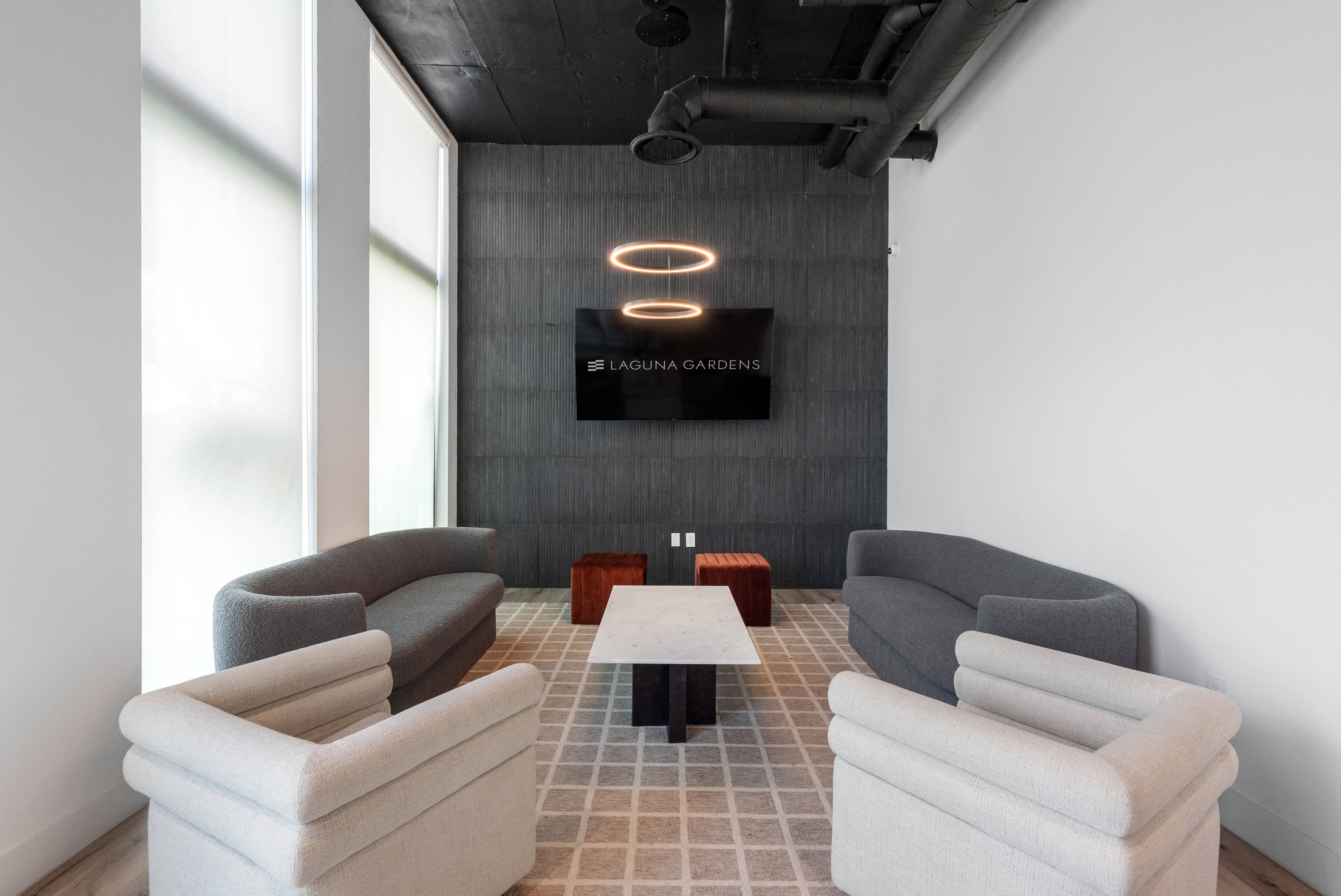 A stylish modern lounge area featuring two curved sofas and two armchairs arranged around a central table, with a wall-mounted screen displaying the text 'Laguna Gardens'.