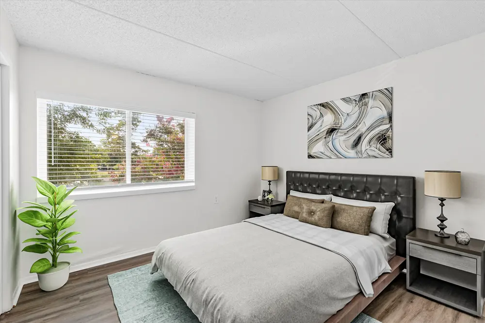 A modern bedroom featuring a comfortable bed with light bedding, a dark headboard, two bedside lamps, and a stylish piece of wall art. A large window allows natural light to fill the room, complemented by a potted plant in the corner and wooden floors.