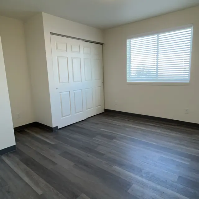 An empty room with gray wooden flooring, a white ceiling light, and a window with white blinds.