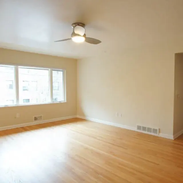 An empty living room with wooden flooring, a ceiling fan, and large windows letting in natural light. A doorway is visible in the corner leading to another space.