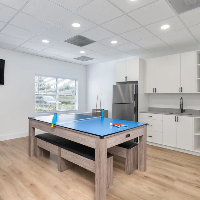 A bright recreational room with a blue table for games, featuring a grey sofa in the corner, a modern kitchen area, and a wall-mounted sign that reads 'VISTA SHORES'.