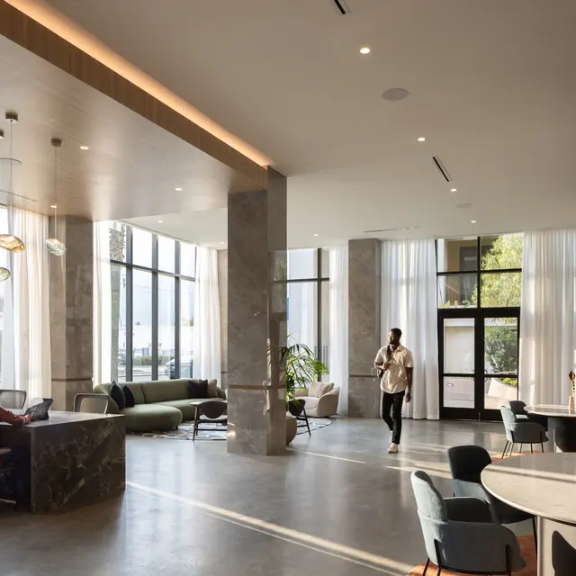 A modern office lobby featuring large windows, stylish furniture, and a workspace. There is a reception desk with a person sitting at it, and another person walking through the space.