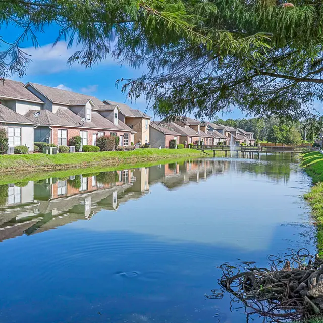 A serene view of a neighborhood alongside a calm water body, featuring reflections of buildings and trees in the water.